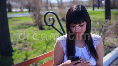 东方美丽的女孩穿着一件白色衬衫<strong>坐在公园里</strong>，在电话上写短信，情感
